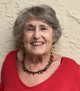 Smiling elderly woman wearing a red top and beaded necklace.