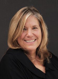 Smiling woman with blonde hair in a black top against a dark background.