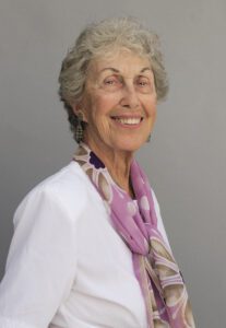 Smiling elderly woman with gray hair wearing a white top and purple scarf.