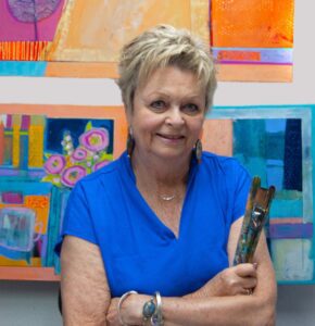 Smiling woman in blue shirt holding a paintbrush in front of colorful artwork.