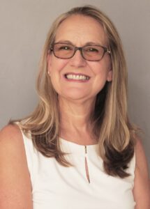 Smiling woman with glasses and light hair wearing a white top.