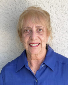Smiling elderly woman in a blue shirt against a white textured wall.