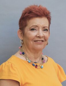 Smiling older woman with short red hair wearing a yellow top and colorful necklace.