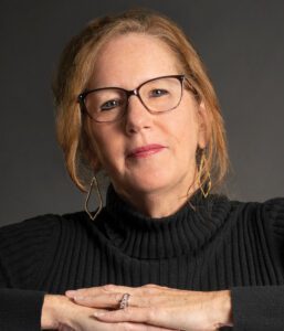 Confident woman with glasses and a black sweater against a dark background.