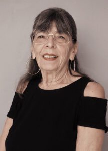 Smiling elderly woman with glasses and hoop earrings in a black top.