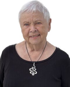 Smiling elderly woman with short white hair wearing a black top and snake-shaped necklace.