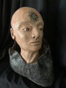 A realistic bust sculpture of a bald figure with a sun tattoo on the forehead.