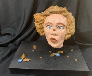 Sculpted blonde head with detailed features on black base with small objects.