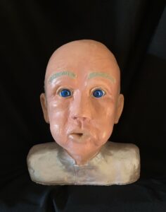A ceramic or plaster bust of a bald man with blue eyes against a black background.