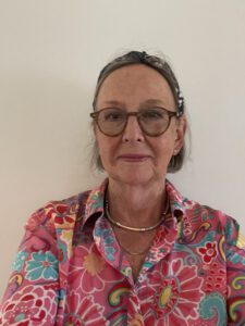 A woman wearing glasses and a colorful floral shirt stands against a plain wall.