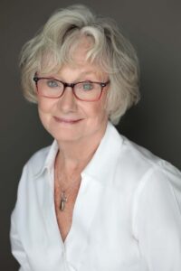 Smiling elderly woman wearing glasses and a white blouse against a dark background.