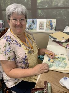 Smiling elderly woman painting detailed bird illustrations at a table.