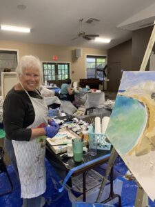 Elderly woman painting a beach scene in an art studio.