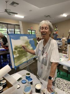 Elderly woman painting a landscape indoors, smiling joyfully.
