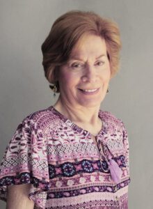 Smiling elderly woman in a patterned top against a plain background.