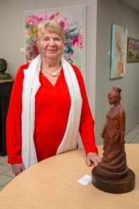 An elderly woman in a red outfit stands beside a wooden sculpture.