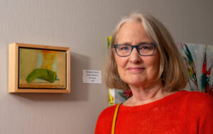 A smiling woman stands beside a small framed artwork on a wall.