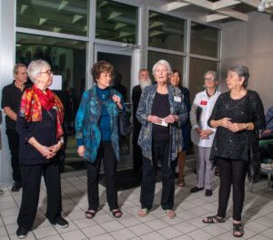 A group of older women standing indoors, engaged in conversation.