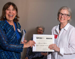 Two women smiling while holding a certificate together indoors.