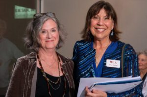 Two smiling women posing indoors, one holding documents.