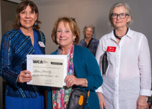 Three women posing together, one holding a certificate from WCAI.