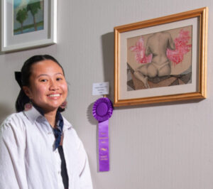 A smiling woman stands beside a framed floral art with a purple award ribbon.