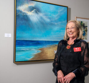 A smiling woman stands beside a vibrant ocean painting in a gallery.