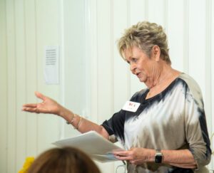An older woman speaking and gesturing while holding papers during a presentation.