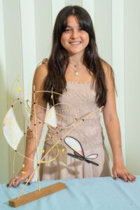 Smiling woman in a light dress standing behind a decorative tree with golden ornaments.