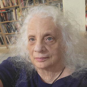 Elderly woman with white curly hair in a cozy room with bookshelves.