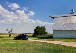 A blue car parked near a white barn on a sunny day with clouds.
