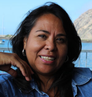 Smiling woman with dark hair by the water and rocky shore in the background.
