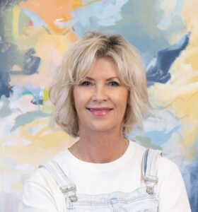 A smiling woman with blonde hair in front of a colorful abstract background.