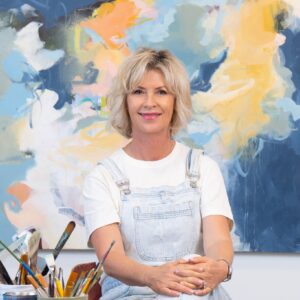 A smiling woman in white overalls poses in front of a colorful abstract painting.