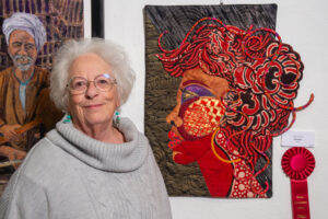 Elderly woman standing next to vibrant textile art of a red-haired woman in profile.