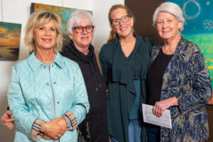 Four smiling women standing together indoors, dressed in casual to semi-formal attire.