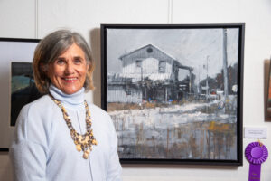 Woman standing beside her abstract painting of a house and landscape.
