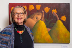 An elderly woman smiling in front of a colorful painting with tulips and abstract shapes.