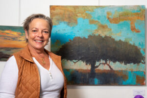 Smiling woman standing beside a colorful tree painting on the wall.