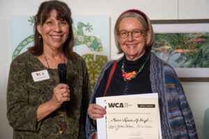 Two women smiling, one holding a microphone and the other a certificate.