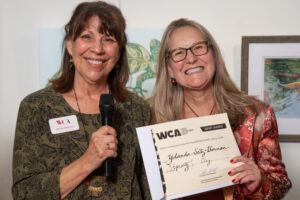 Two women smiling, one holding a microphone, the other holding a certificate from WCA.