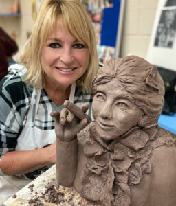 Woman smiling behind a detailed clay sculpture of a figure blowing a kiss.