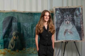 A woman with long hair stands beside an artwork on an easel.