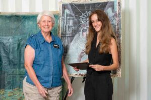 Two women smiling in front of artwork, one holding a palette.