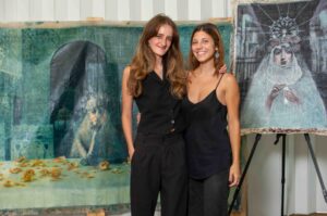 Two women posing together in front of artworks, dressed in black.