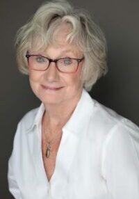 Smiling elderly woman wearing glasses and a white blouse against a dark background.